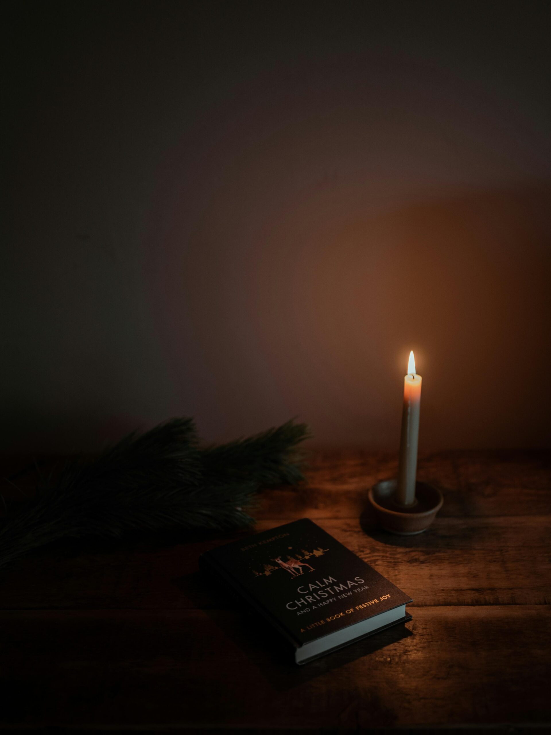 A lit candle in a small holder sits on a wooden table beside a book titled Calm Christmas and some pine branches, creating a warm and cozy atmosphere in dim lighting.