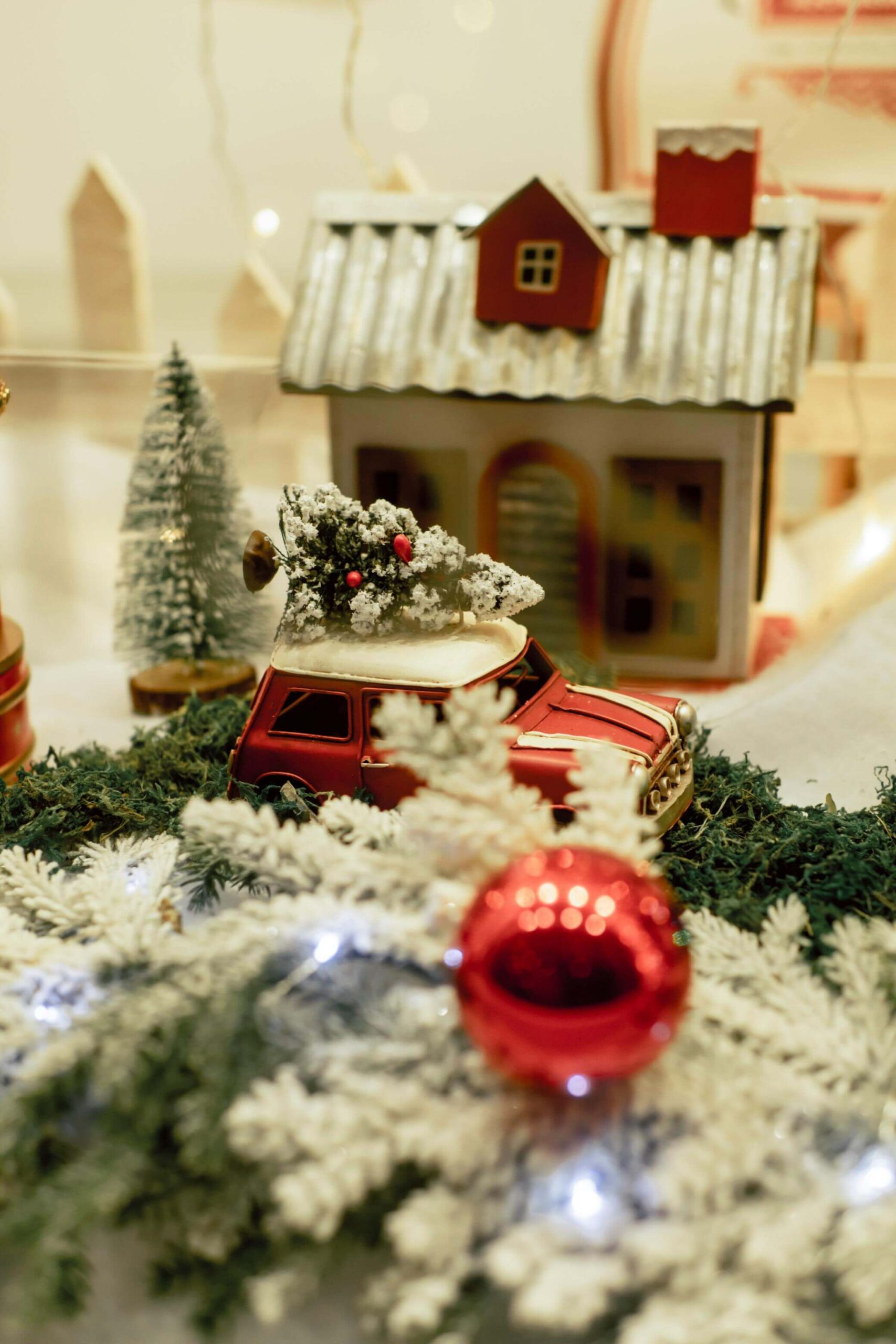 A miniature red car with a snow-covered Christmas tree on its roof sits in front of a small decorative house, surrounded by fake snow, a red ornament, and festive greenery.
