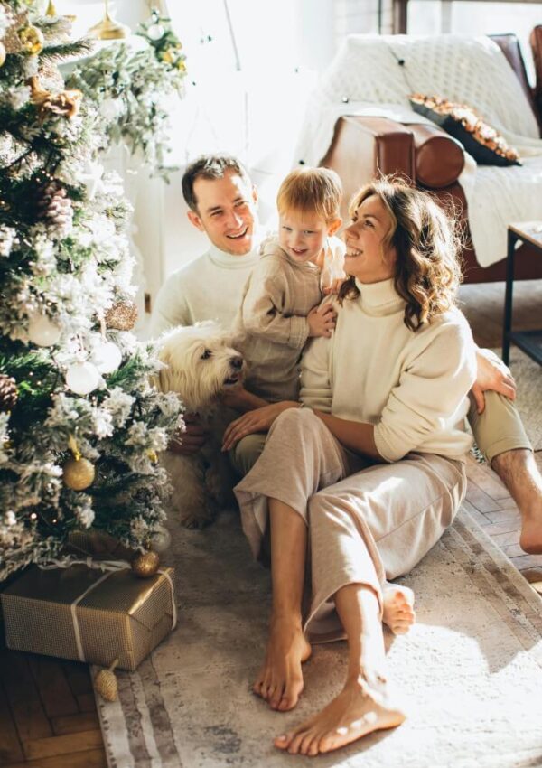 A smiling family sits together by a decorated Christmas tree with wrapped gifts. A woman, man, child, and fluffy white dog are bathed in warm sunlight in a cozy, festive living room.