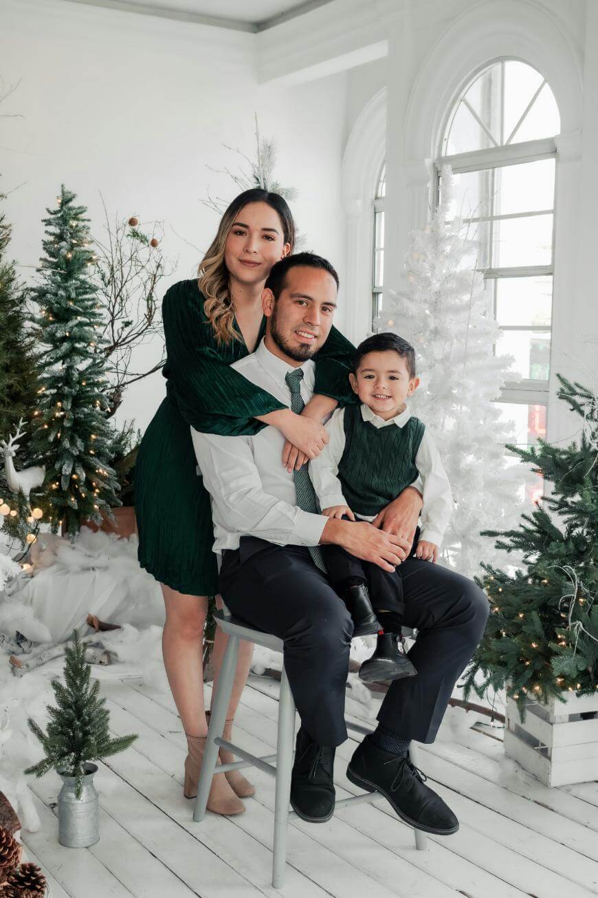A family poses for a holiday photo in a decorated room with Christmas trees. A woman stands behind a seated man, both hugging a young boy on the man’s lap. All are wearing coordinated green and white outfits.