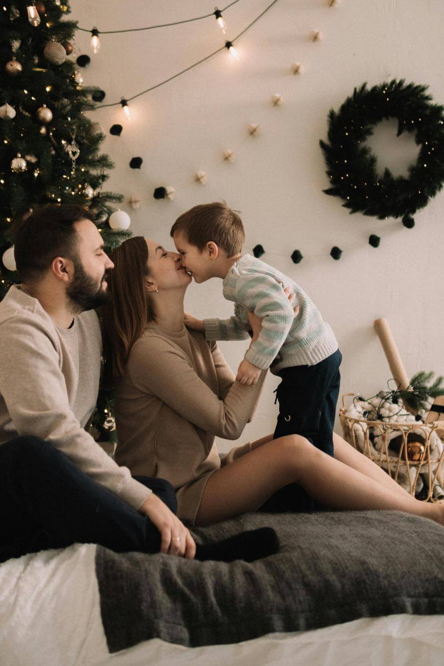 A family sits by a Christmas tree and wreath; a woman lifts a young boy who kisses her nose, while a man sits beside them, all smiling and surrounded by festive decorations.