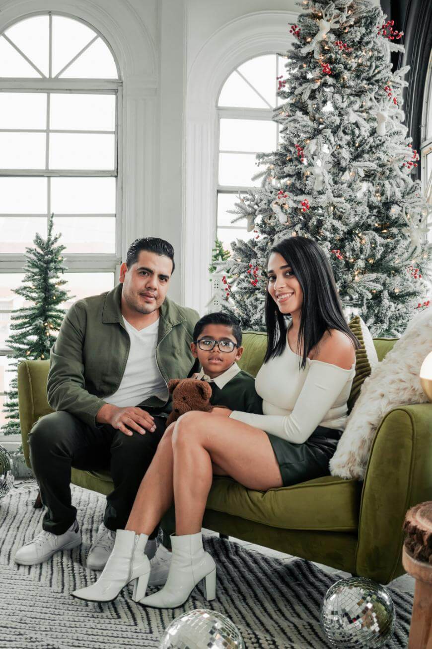 A family of three poses indoors by a decorated Christmas tree. The man and woman sit on a green sofa with a young boy holding a stuffed animal between them. Large windows and festive decor are in the background.