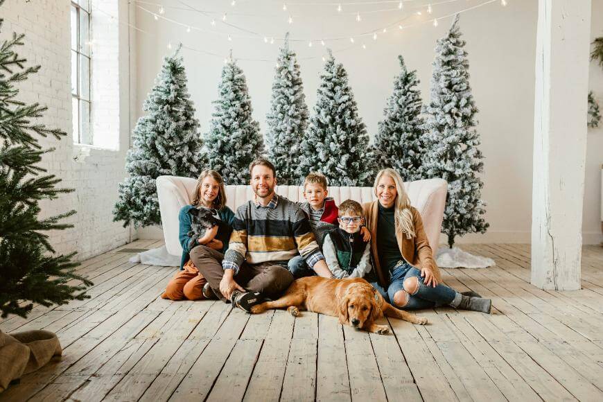 A family of five, including two adults, three children, and a dog, sits smiling on a wooden floor in front of snowy Christmas trees and string lights in a cozy, festive setting.