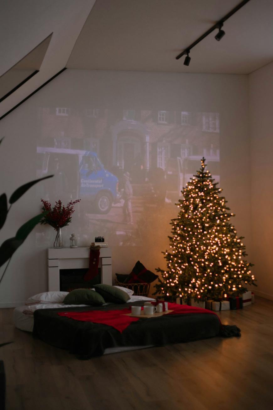 A cozy living room with a lit Christmas tree, a mattress on the floor with red and black bedding, and a movie projected on the wall. Presents are under the tree, and soft lighting creates a warm, festive atmosphere.