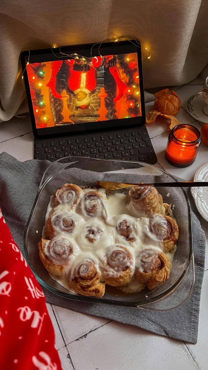 A glass dish of frosted cinnamon rolls sits on a gray cloth next to a red candle, an orange, and a tablet playing a Christmas movie with festive lights. A red blanket with white text is partially visible.