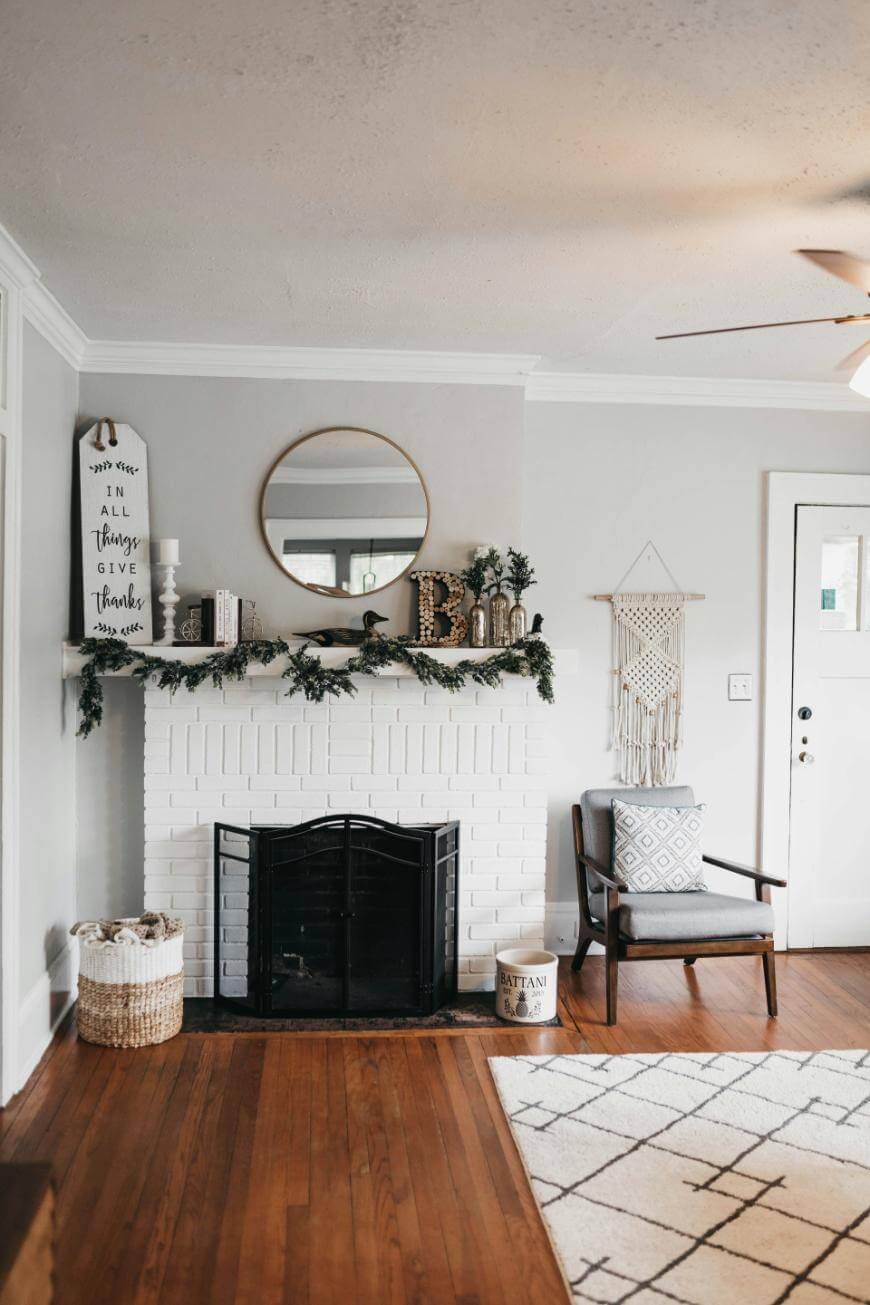 A cozy living room with a white brick fireplace decorated with garland, books, and candles, a round mirror above, a woven basket, an armchair with a patterned cushion, and a macramé wall hanging on a light gray wall.