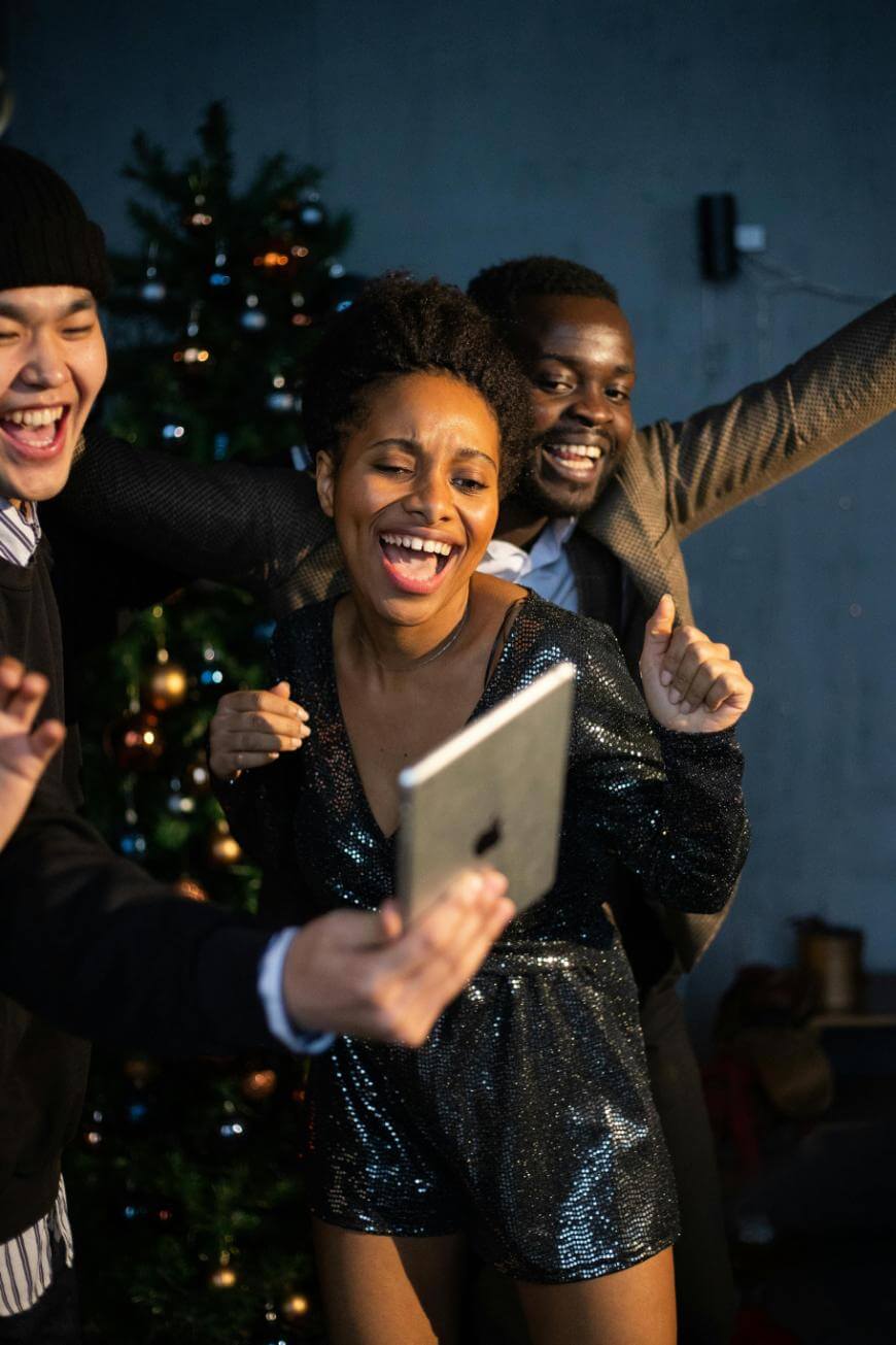 Three friends are smiling and dancing together in front of a decorated Christmas tree, posing for a selfie with a tablet. The atmosphere is joyful and festive.
