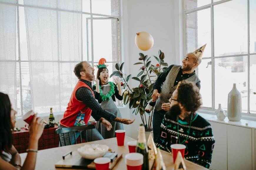 A group of people in festive clothing laugh and play with a balloon at a holiday party. They wear party hats and hold red cups, surrounded by holiday decorations and natural light from large windows.