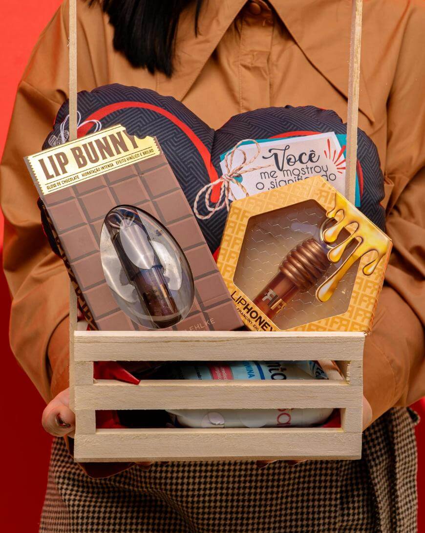 A person in a brown shirt holds a wooden crate containing a heart-shaped pillow, a Lip Bunny lipstick, a Lip Honey lip balm shaped like a honey dipper, and other gift items against a red background.