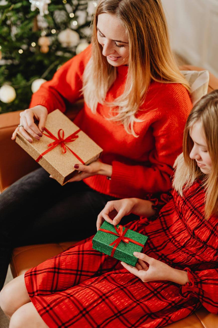 Two women in red dresses sit by a Christmas tree, holding wrapped presents with red ribbons, smiling and preparing to exchange gifts.