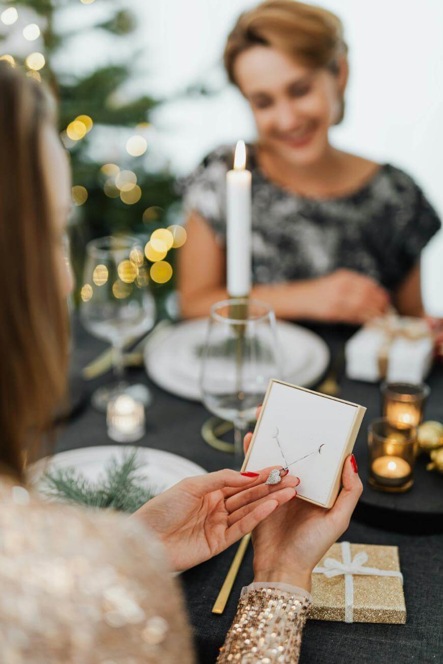 A person holds an open jewelry box with a necklace at a festive dinner table, while another person sits and smiles in the background with holiday decorations and candles around them.
