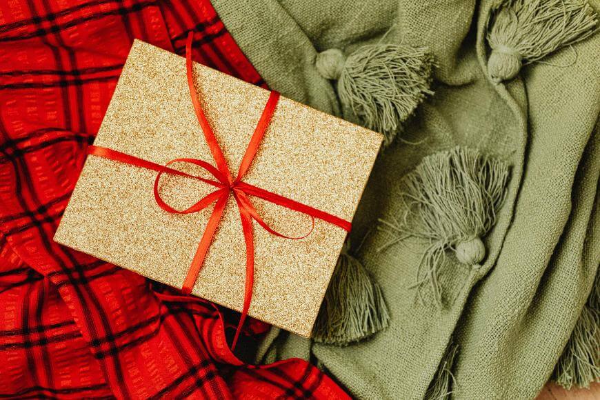 A gold glittery gift box with a red ribbon sits on a green tasseled blanket and a red plaid fabric.