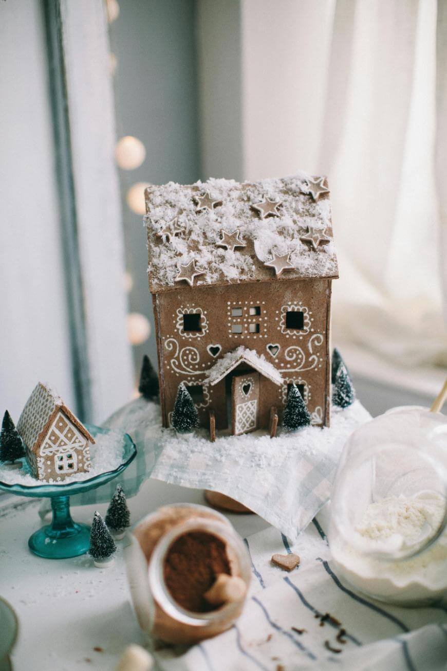 A large gingerbread house decorated with white icing and powdered sugar sits beside a smaller gingerbread house on a blue stand, surrounded by miniature trees and baking supplies on a light-colored table.