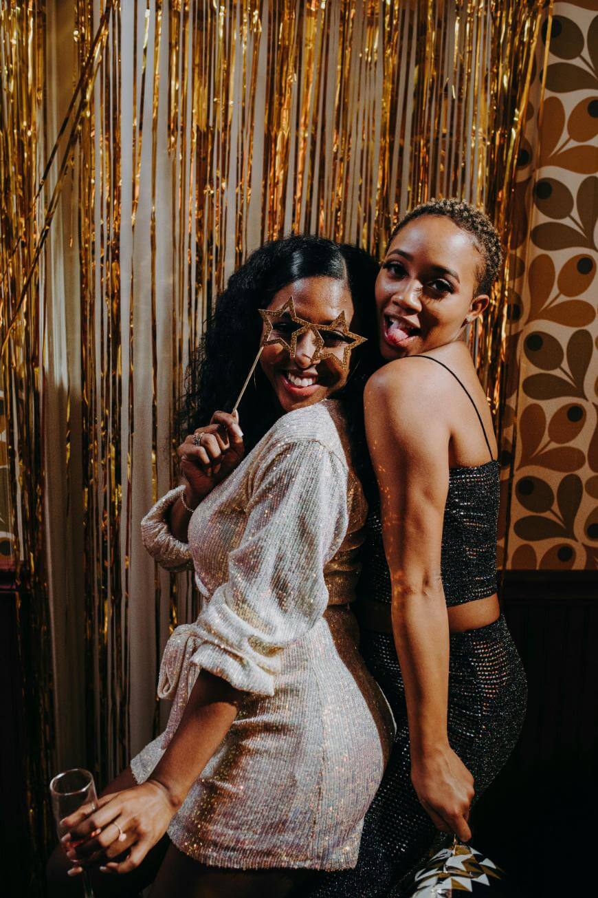 Two women in sparkly outfits smile and pose playfully at a party, one holding star-shaped glasses to her face. Gold streamers and retro-patterned wallpaper create a festive background.
