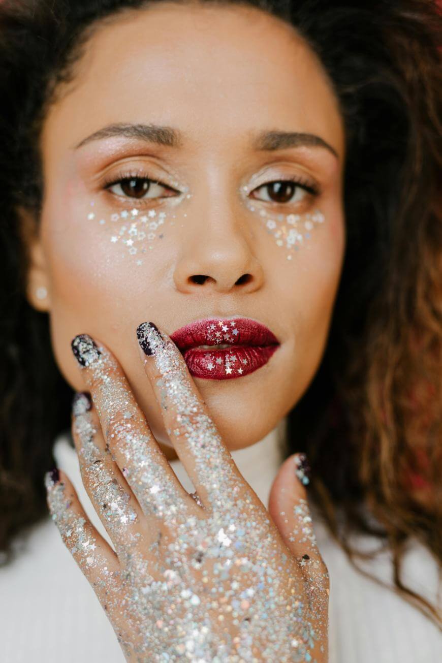 A person with curly hair and bold red lipstick has silver glitter and star-shaped sequins on their face and hand, creating a sparkling, festive look. They gently touch their lips with their glitter-covered fingers.