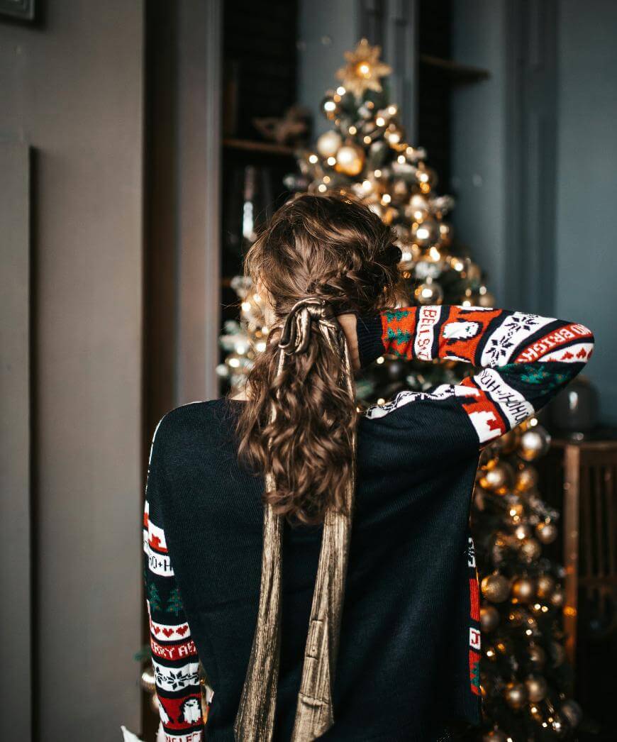 A person with curly hair tied with a gold ribbon stands facing a decorated Christmas tree, wearing a festive sweater with holiday patterns, in a warmly lit room.