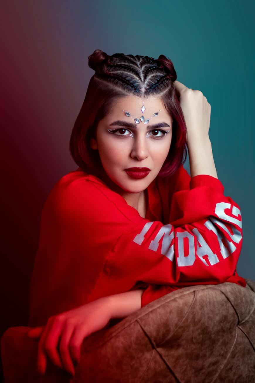 A woman with braided hair styled in two buns, wearing a red sweatshirt with white text on the sleeve, sits on a sofa. She has decorative gems on her forehead and dramatic makeup, looking confidently at the camera.