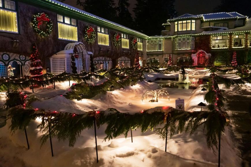 A large house decorated with Christmas lights and wreaths, with snow-covered gardens bordered by greenery and lights, creating a festive and cozy winter holiday scene at night.