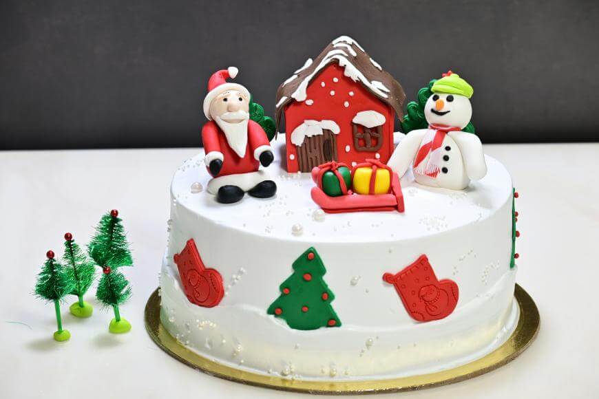 A white Christmas cake decorated with a Santa figurine, a snowman, a red house, and presents on top. The sides have red and green festive shapes, and there are three small model Christmas trees beside the cake.