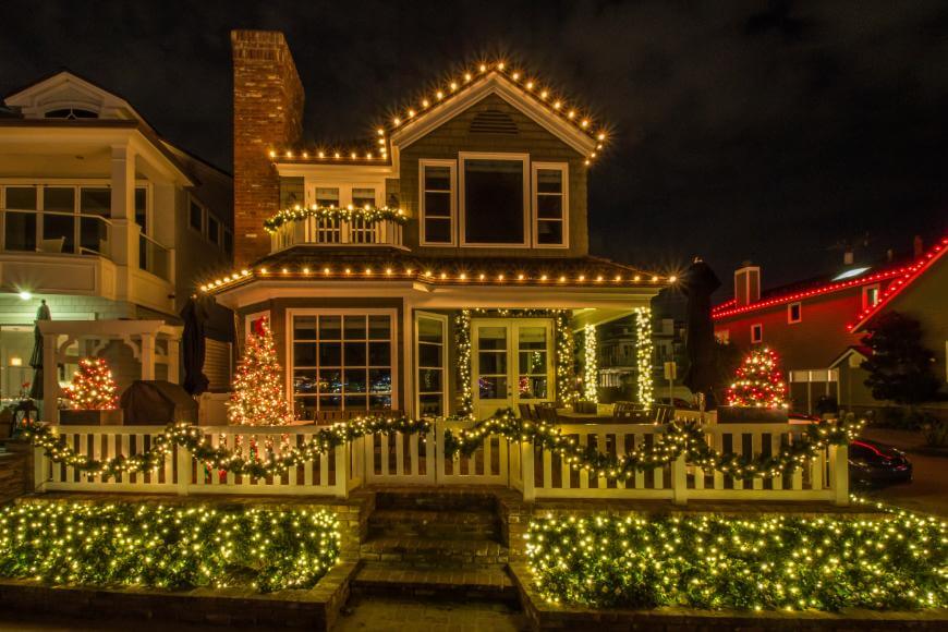 A two-story house is decorated with yellow Christmas lights along the roof, porch, and railing. Two lit Christmas trees sit by the entrance, and other houses with festive lights are visible nearby at night.