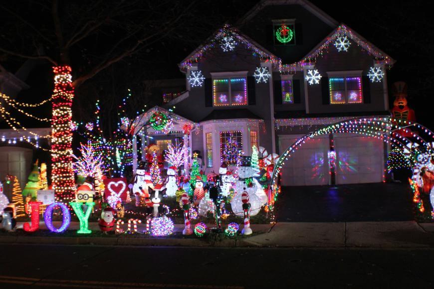 A house and yard are covered in colorful Christmas lights and decorations, including snowmen, reindeer, and the word JOY. Bright lights and festive ornaments fill the scene, creating a cheerful holiday display at night.