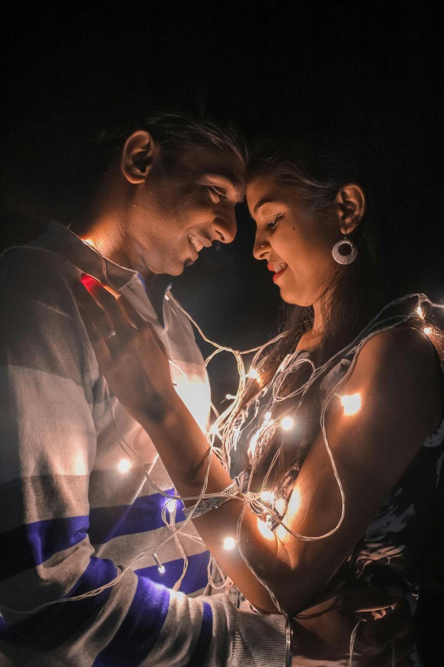 A couple stands close together, smiling and gazing into each others eyes, wrapped in glowing string lights against a dark background.