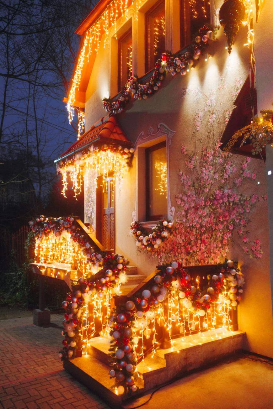 A house decorated with warm white string lights and garlands featuring red, silver, and gold ornaments wraps around the balcony, railings, and windows. Pink floral wall art is visible on the exterior near the entrance.