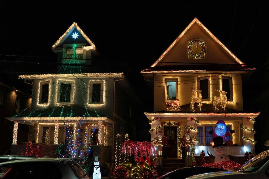 Two neighboring houses decorated with colorful Christmas lights at night. One house has lights forming a tree shape; the other has reindeer, a wreath, and a large inflatable snowman on the porch. Cars are parked in front.