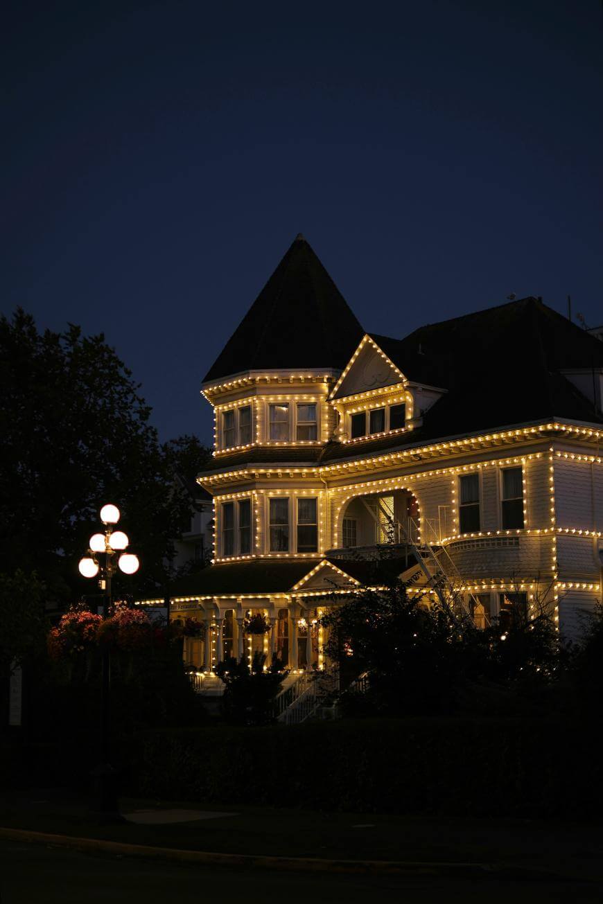 A large, Victorian-style house is outlined with warm white string lights at dusk. A vintage streetlamp glows in the foreground, and the house is surrounded by dark trees and shrubs.