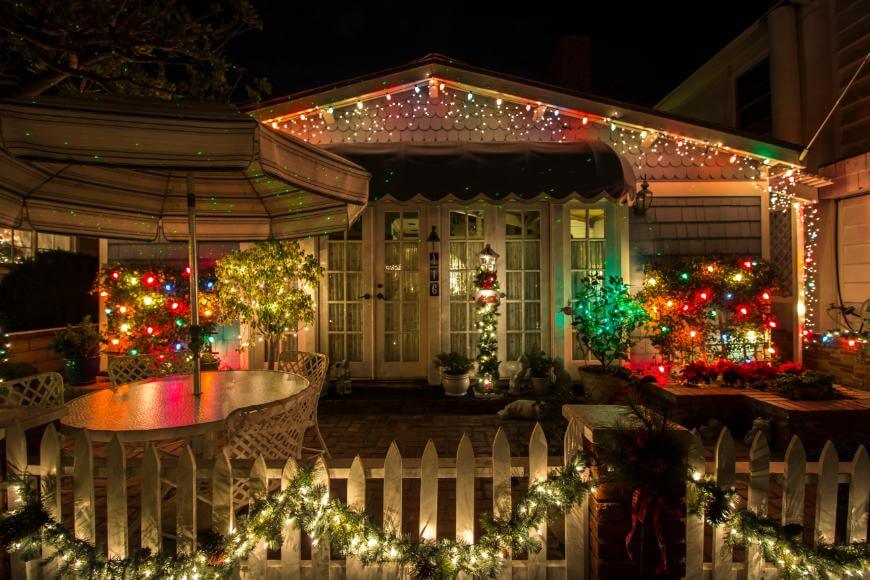 A cozy house at night is decorated with colorful Christmas lights and garlands. The porch is glowing, a white fence is wrapped in lights, and a patio table sits under an umbrella in the festive scene.