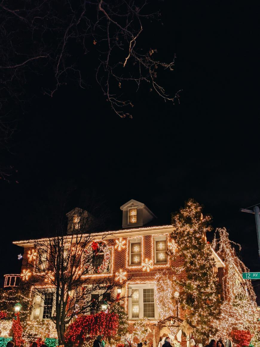 A brick house decorated with numerous white and yellow Christmas lights, with illuminated trees and wreaths, glowing warmly at night against a dark sky. A green street sign reads “12th AV.”.