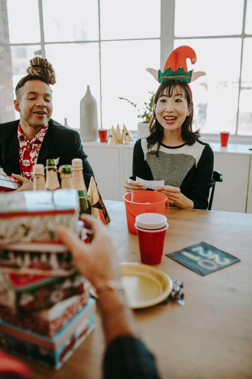 Two people sit at a table with drinks, red cups, and holiday decorations. One person wears an elf hat and holds cards, smiling, while others participate in a festive gathering.