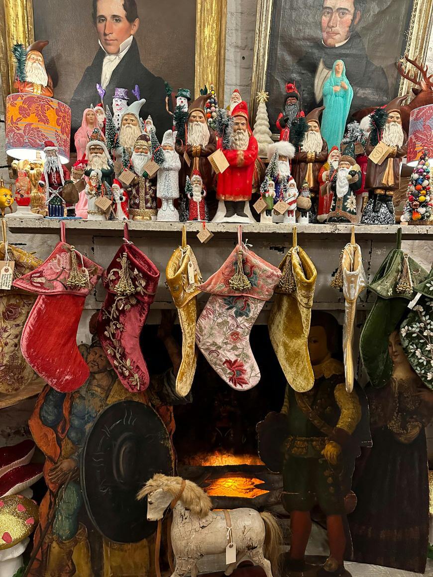 A decorated mantel displays vintage Santa figurines, Christmas stockings, and portraits. Below, a toy horse, cardboard cutouts, and a glowing fireplace create a festive, nostalgic holiday scene.