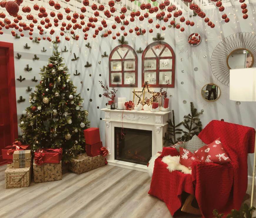 A cozy Christmas living room with a decorated tree, wrapped gifts, a white fireplace with candles and ornaments, a red armchair with a throw and cushion, and red baubles hanging from the ceiling.