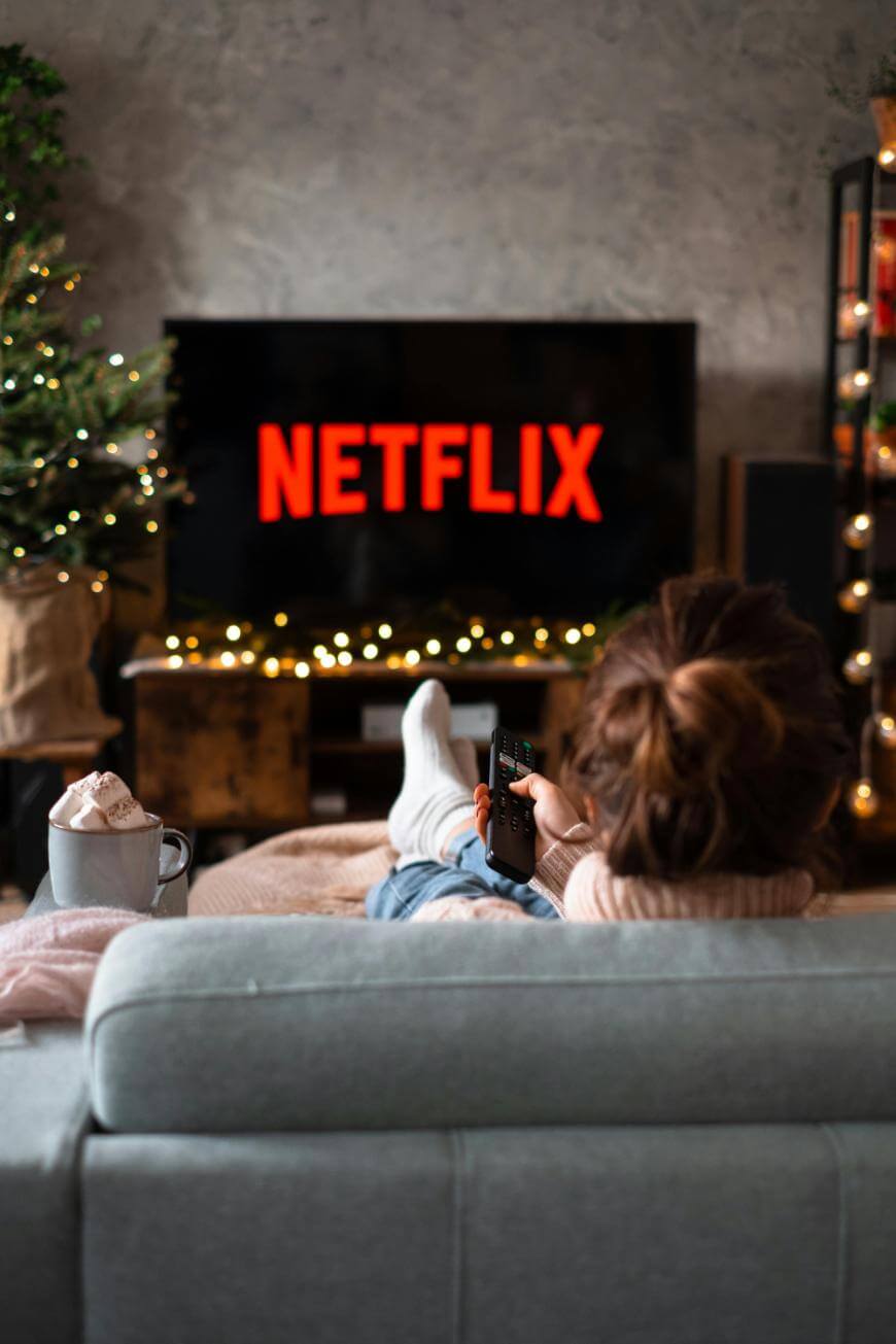 A person with brown hair in a bun relaxes on a couch with a blanket and a hot drink, watching Netflix on a large TV with holiday lights and a decorated tree in the background.
