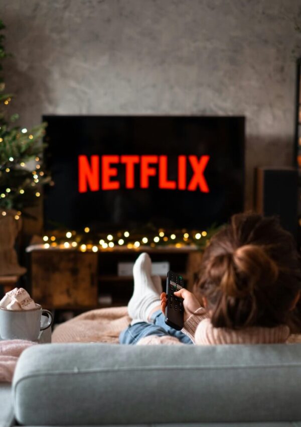 A person sits on a couch with a remote control, facing a TV displaying the Netflix logo. The room is decorated with string lights and a Christmas tree, and there is a mug of hot chocolate on the table.