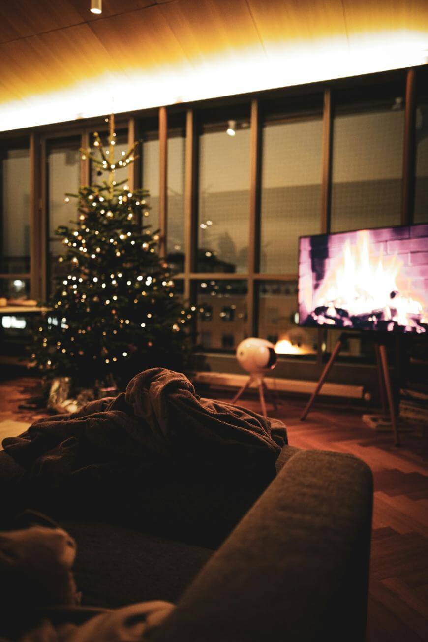 A cozy living room with a lit Christmas tree, a TV displaying a fireplace, and a sofa with a blanket, softly illuminated by warm lights from the ceiling and decorations.