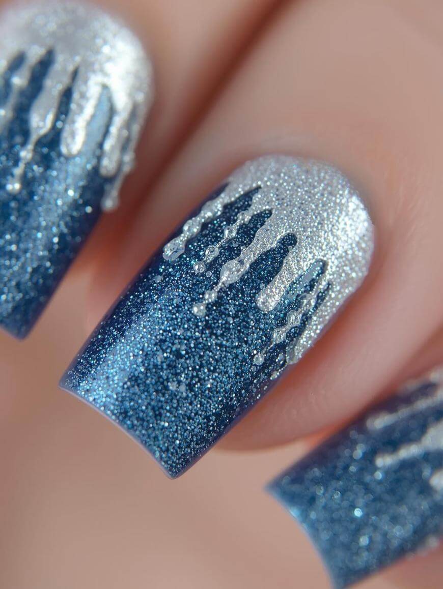 Close-up of fingernails with blue glittery polish, topped with silver glitter resembling dripping icicles or melting snow for a winter-themed nail art design.
