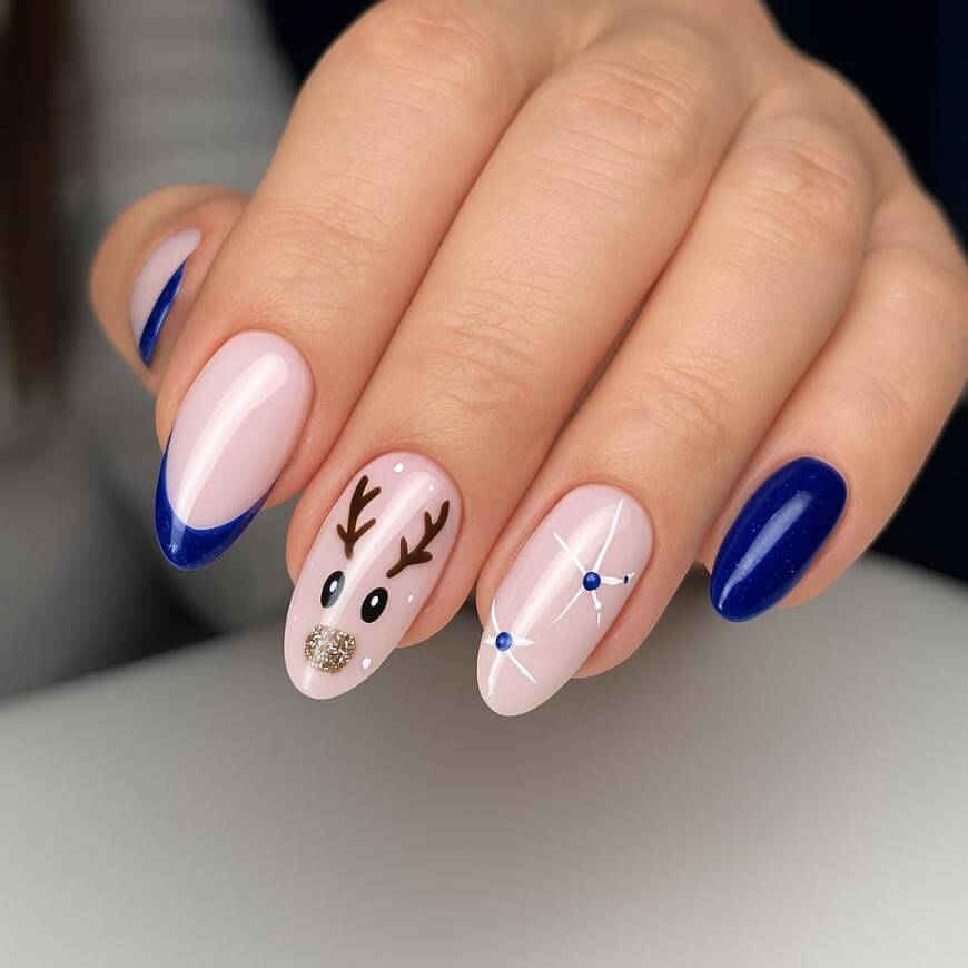 A hand with manicured nails featuring blue French tips, solid dark blue polish, a reindeer face design on the middle finger, and white with blue star-like dots on the ring finger.
