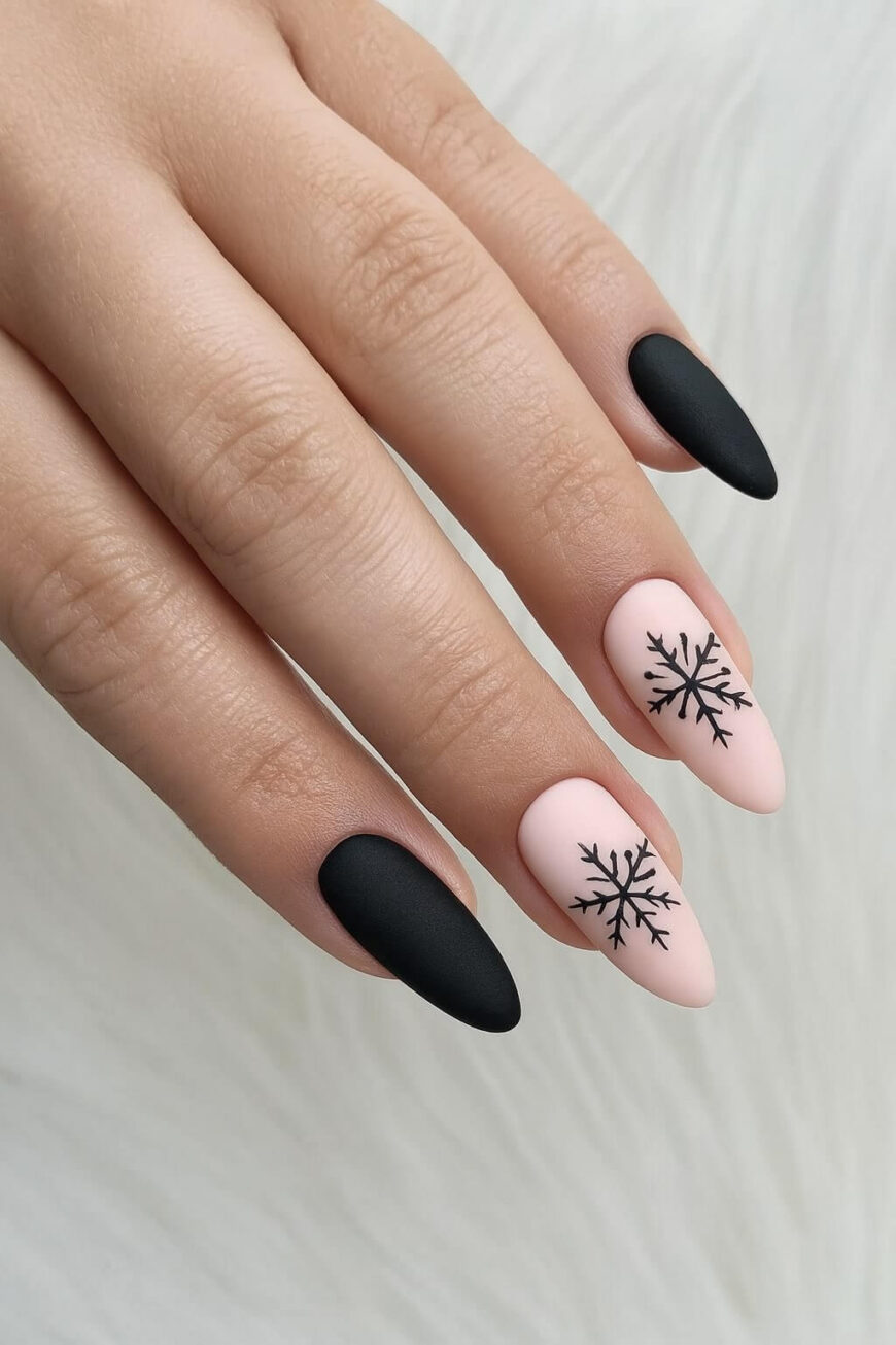 A hand with almond-shaped nails featuring a matte black polish on the thumb, index, and pinky, and light pink polish with black snowflake designs on the middle and ring fingers, resting on a white surface.
