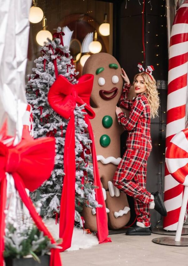 A woman in red plaid pajamas and reindeer antlers poses next to a large gingerbread man decoration, festive Christmas trees, oversized candy canes, and big red bows outside a holiday-themed storefront.
