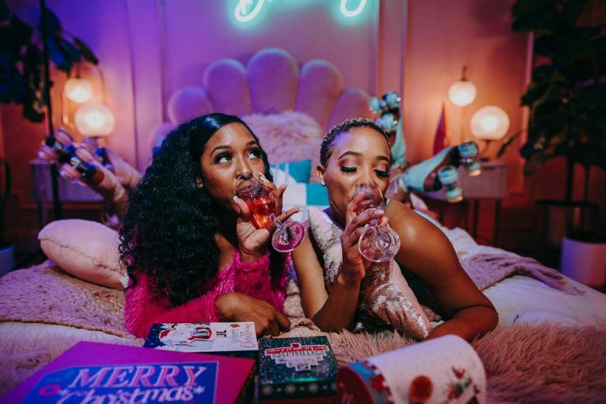 Two women in festive outfits lay on a cozy bed, sipping drinks from wine glasses. Around them are Christmas-themed gift boxes and decorations, creating a warm, celebratory atmosphere.