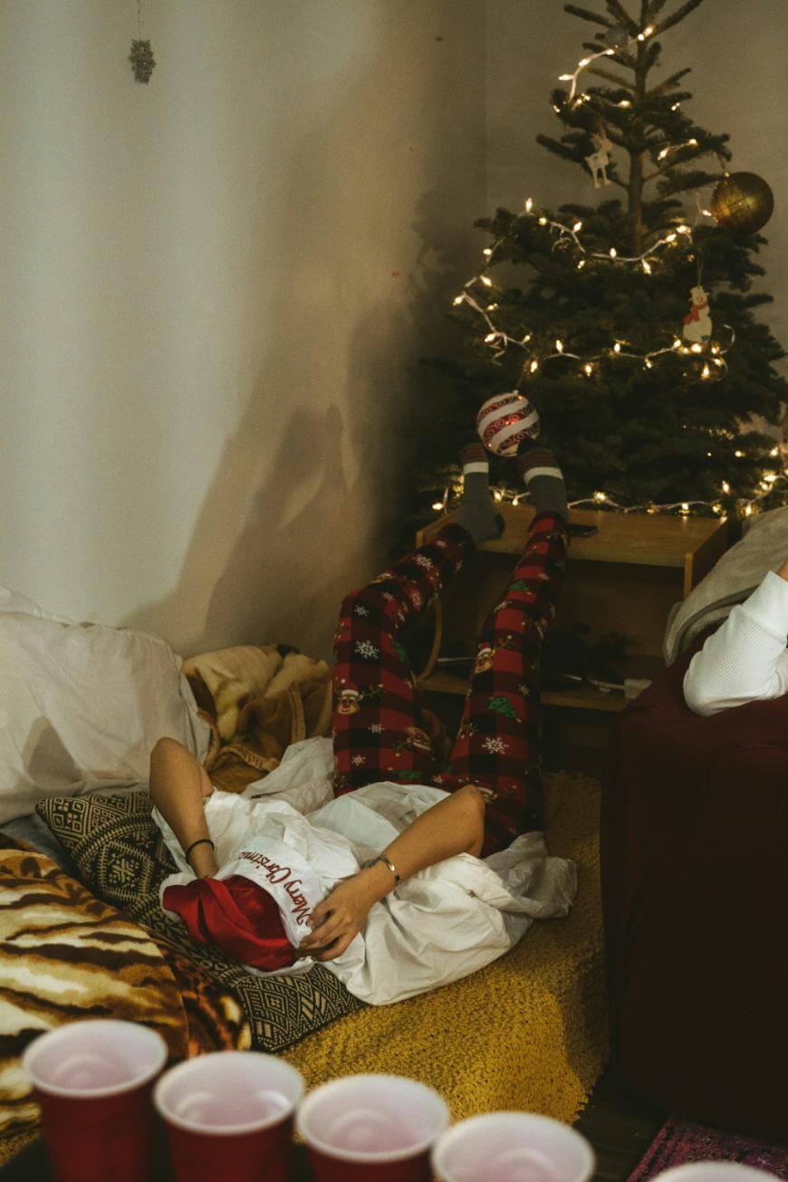 A person in festive pajamas and a Santa hat lies on a couch with legs up against a wall, near a lit Christmas tree. Red cups are visible in the foreground, and blankets are scattered around.