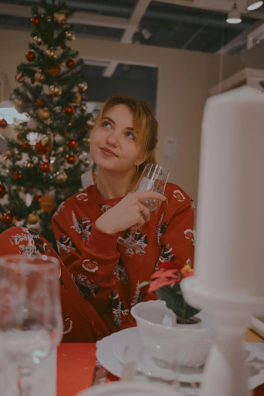 A woman in red festive pajamas sits in front of a decorated Christmas tree, holding a wine glass and smiling thoughtfully. There are plates, glasses, and a small poinsettia on the table in the foreground.