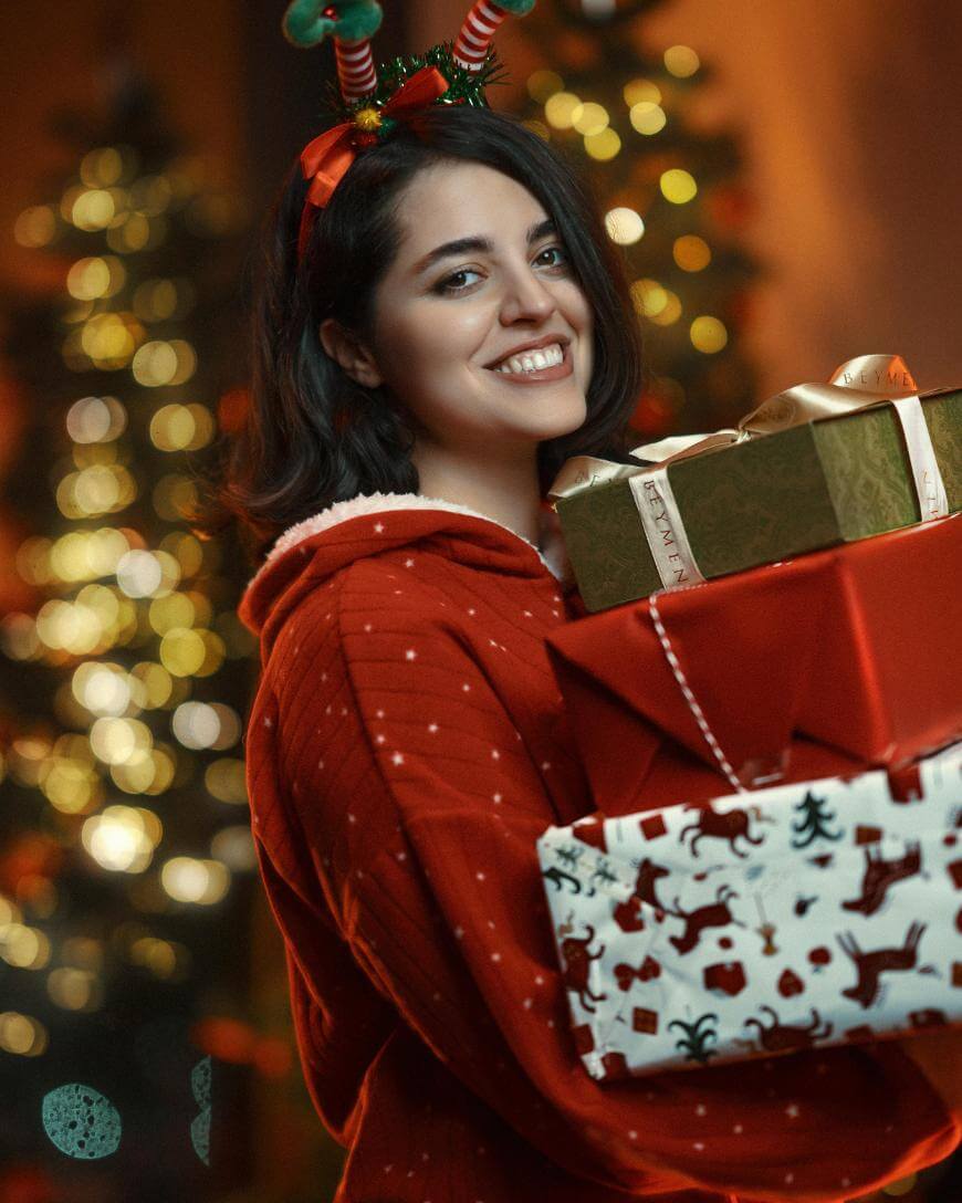 A smiling woman in a red festive outfit and reindeer antler headband holds three wrapped Christmas gifts, with decorated Christmas trees glowing in the blurred background.
