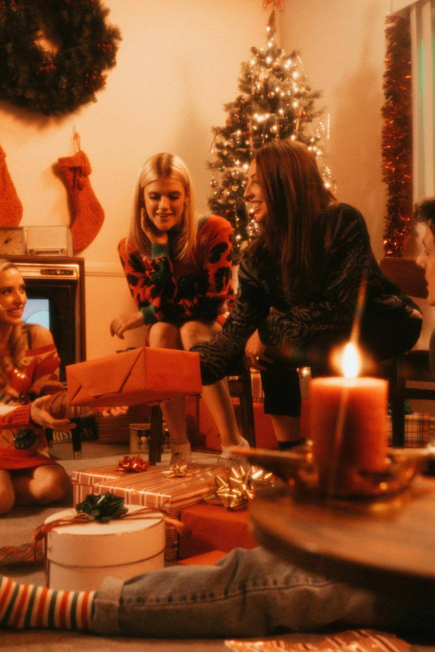 A group of friends in festive sweaters gather around a Christmas tree, exchanging gifts and smiling. The room is decorated with stockings, a wreath, and presents, with a lit candle in the foreground.