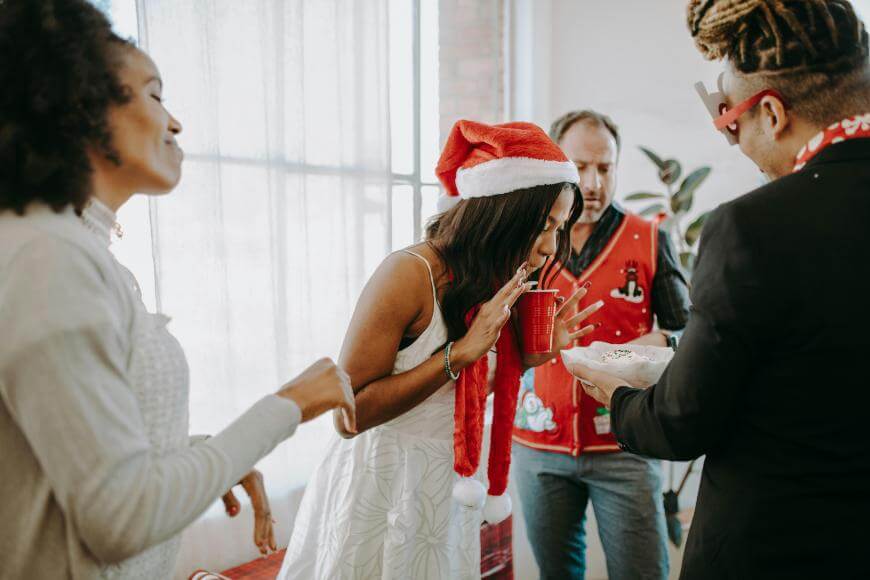 Four people at a festive gathering; one woman in a Santa hat leans in excitedly toward a plate held by a man in reindeer glasses. Another woman and man stand nearby, all appearing cheerful and engaged.