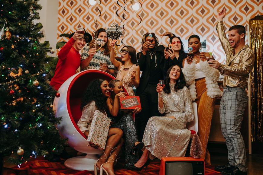A group of people in festive, retro clothing gather near a decorated Christmas tree, raising glasses and celebrating in a room with bold patterned wallpaper. Some are sitting, others standing, all appearing joyful and lively.