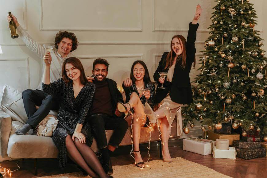 Five people dressed in festive attire sit and stand around a decorated Christmas tree, smiling and raising drinks, celebrating together in a warmly lit living room with wrapped gifts nearby.