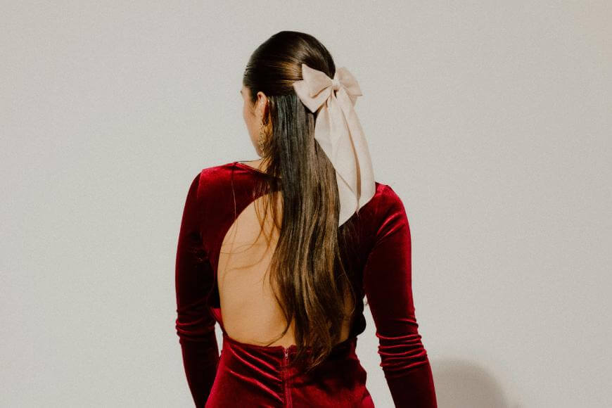A woman with long brown hair, wearing a large beige bow, is seen from behind in a backless red velvet dress against a plain light background.